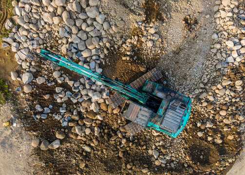 Aerial view of a teal excavator working on a construction site surrounded by large rocks and gravel in Karanglewas, Central Java, Indonesia.