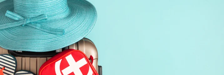First aid kit, blue straw hat, and flip flops on suitcase against turquoise background with copy space. Concept of safe summer vacation, medical insurance for travel, and trip preparation. © prime1001