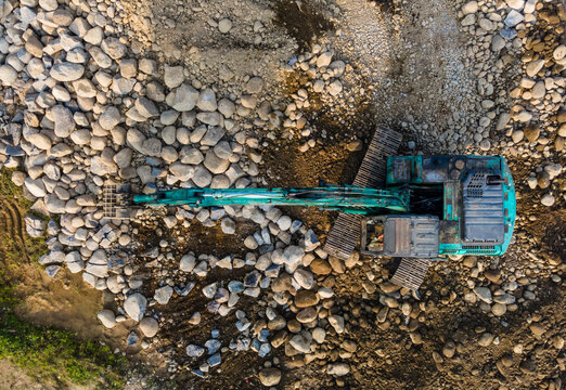 Aerial view of a teal excavator working on a construction site at Babakan surrounded by large river stones and boulders in Karanglewas, Central Java, Indonesia.