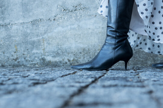 High angle close up of a single black leather ankle boot with high heel on cobblestone street, under a flowing polka dot dress.