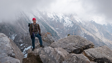 A person stands confidently on large rocks in a mountainous area. The sky is cloudy, and snow can be seen on some peaks. This scene shows an outdoor adventure and exploration. © Evgeniy