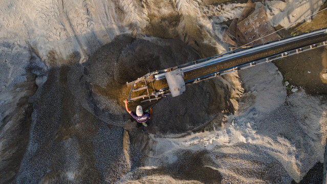 Aerial view of a worker standing by a conveyor belt pouring sand and gravel onto a large pile at a quarry in Karanglewas, Central Java, Indonesia.