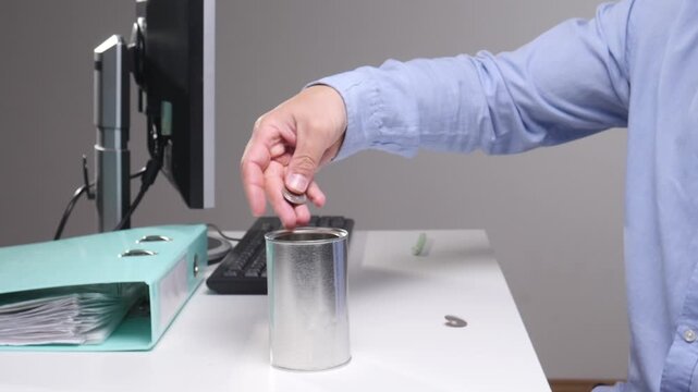 Person's hand dropping a coin into a silver collection can on a white office desk, symbolizing concepts of saving, charity, fundraising, and financial contribution