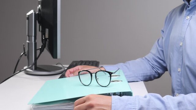 Office worker organizing a stack of documents in a binder while using a computer, with reading glasses resting on top of the file, representing busy professional work and administration