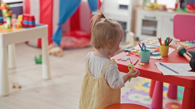 Toddler girl holding scissors at red table playing with art supplies in studio; playfulness imagination discovery.