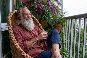 Obraz premium Content elderly man with long white beard seated in wicker chair on balcony holding a cup for wellness wallpaper and home lifestyle design