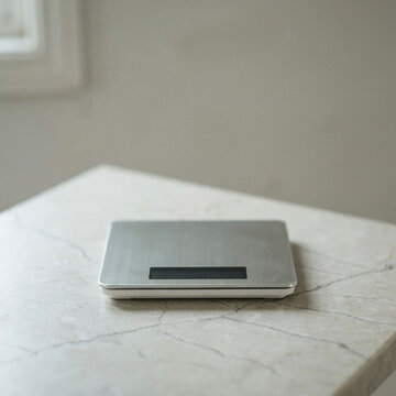 Digital kitchen scale with a brushed metallic finish sits alone on a light stone countertop in a bright interior space.