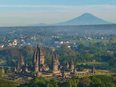 Aerial view of the ancient Prambanan Temple complex with stone spires set against lush greenery and a distant volcano in Kecamatan Prambanan, Daerah Istimewa Yogya, Indonesia.