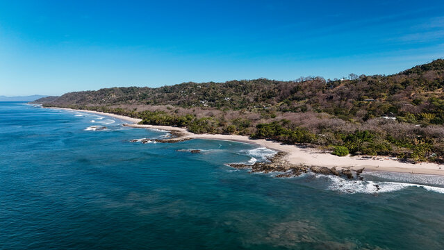 Aerial view of the pristine sandy beach and rocky shoreline of Santa Teresa bordered by lush tropical forest under a clear blue sky in Santa Teresa, Puntarenas Province, Costa Rica.
