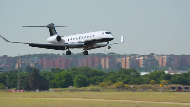 White and black large cabin business jet performs landing at a busy airport with urban cityscape and airport hangar buildings in background, daytime.
