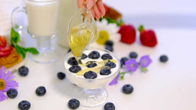 Creamy yogurt is poured into a glass bowl, topped with fresh blueberries and drizzled with golden honey. Bright breakfast scene with chamomile flowers and clean advertising space.