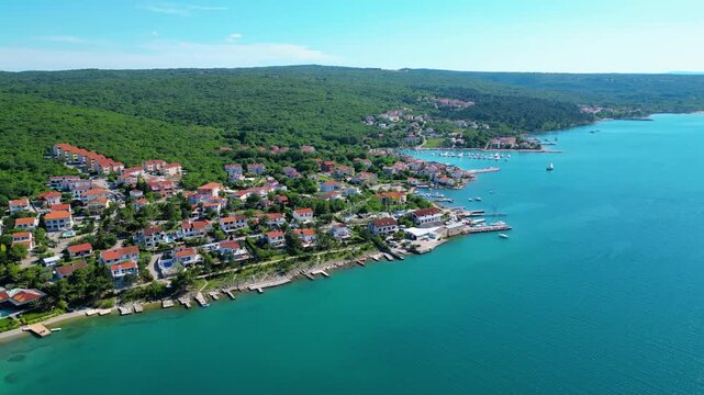 Aerial view of a charming coastal village on Krk Island, Croatia. Turquoise waters meet lush green hills, dotted with red-roofed homes and a bustling marina. Perfect summer escape. Klimno