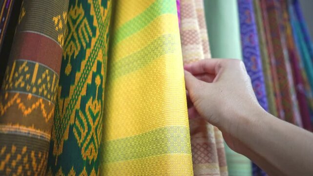 Hand touching rows of colorful traditional Thai silk fabrics, close up , Traditional Isan Thai silk fabrics featuring geometric and elephant patterns.