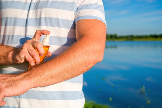 Close up of man spraying tanning oil on arm near lake in summer. Concept of sun care, beach skincare and outdoor relaxation.