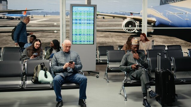 Tourist in airport wearing ethnic clothes using mobile phone to text family about arriving home, African man next to international travelers using smartphone to check flight schedule status, camera A.