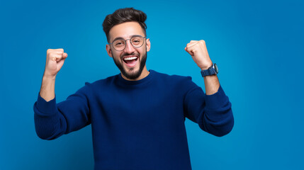 Naklejka premium Confident Excited Young Man Celebrating Success on Blue Studio Background, Happy Winner with Raised Fists, Positive Emotion, Achievement, Business Motivation, Casual Male Portrait