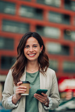 Young businesswoman smiling at camera, holding a hot beverage and mobile phone, standing outdoors during a break