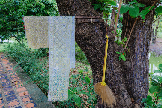 A traditional Thai silk scarf and a broom hang on a tree in Ayutthaya Historical Park, Ayutthaya, Thailand, near a pond, suggesting preparation for a ceremony or daily chores.