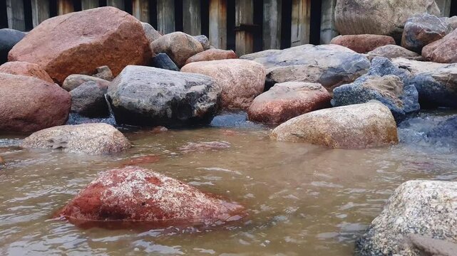 Close-up of flowing clean shallow river or water stream with sand bottom among huge boulders on rocky shore of Baltic sea. Abstract minimalist aquatic texture pattern natural background.