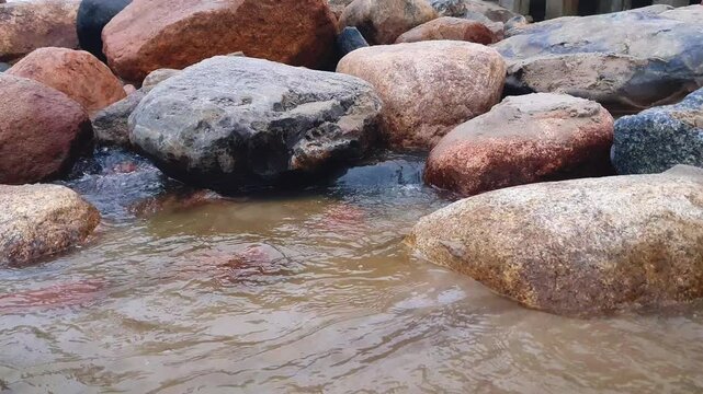 Close-up clean wellspring of flowing water stream or shallow river with sand bottom among huge boulders on rocky shore of Baltic sea. Abstract minimalist aquatic texture pattern natural background.