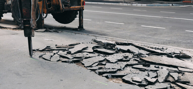 Heavy machinery with a hydraulic hammer breaking up asphalt pavement, performing road construction and repair work on a city street
