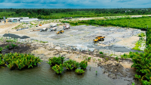 Aerial view of a construction site with cement mixer trucks, heavy machinery, and concrete foundations near a river and lush tropical vegetation in Rivers State, Nigeria.