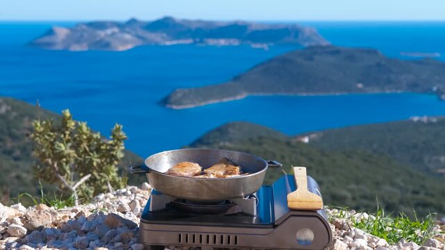 Cooking meal on portable stove with stunning sea view. Meat frying in a pan on a portable camping stove with a beautiful scenic background of blue sea and green islands. Outdoor cooking concept