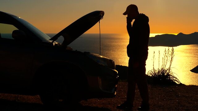 Stranded man calling for help with broken car at sunset. Silhouetted man standing near a broken-down car with an open hood, talking on a mobile phone against a stunning sunset over the ocean