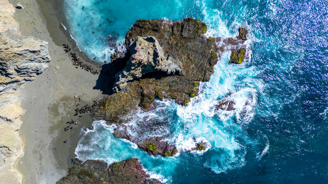 Aerial view of a colony of sea lions resting on a sandy beach beside turquoise ocean waves crashing against rugged rock formations in Magdalena Bay, Mexico.