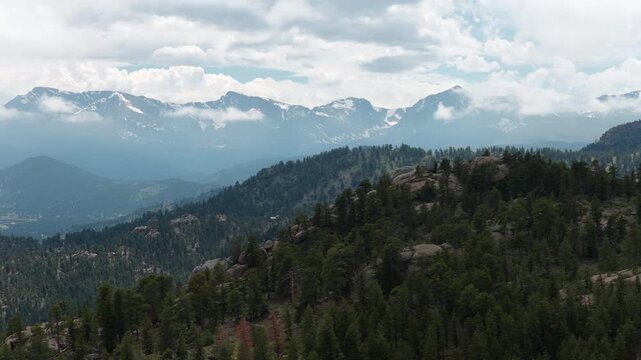 Summer aerial of Rocky Mountain National Park and forest in Estes Park with dense evergreen trees in the foreground and multiple layers of snowcapped peaks under a cloudy sky