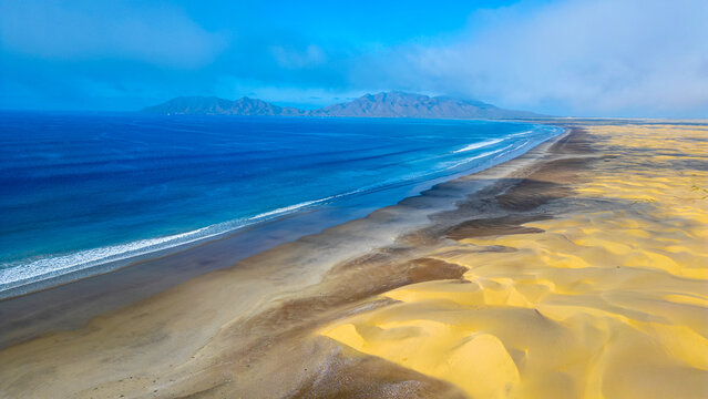 Aerial view of the vast golden sand dunes of Magdalena Bay meeting the deep blue Pacific Ocean with mountains in the background Mexico.