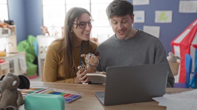 Woman holds giraffe puppet with hand and man watches laptop in preschool classroom; joyful learning.