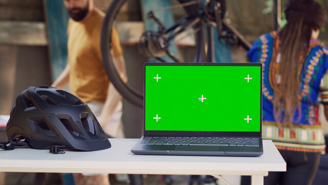 Laptop with green screen display placed near helmet while interracial couple performs bike maintenance in background, with in yard minicomputer is on table showing isolated chromakey template.