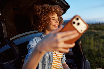 Smiling woman with curly hair taking a selfie in an open car trunk on a sunny day, capturing a casual lifestyle moment and joyful self expression. With natural light and a bright open background. © SHOTPRIME STUDIO