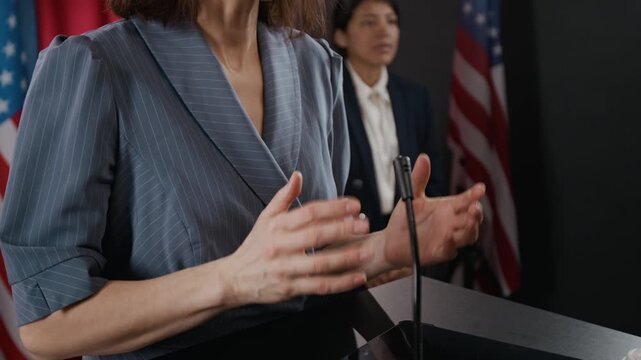 Adult woman gesturing while speaking at podium during press conference with USA flags. Clip is ideal for news broadcasts, political campaigns, and public relations videos, close up shot
