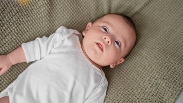 Baby newborn hispanic boy lying on knitted blanket on bed in studio, wearing white onesie, gazing upward with visible hand and open eyes; calm curiosity.