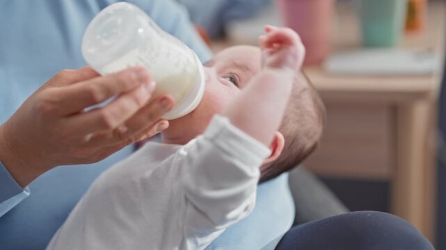 Baby boy fed from bottle by adult hand, cheek and ear visible, held in lap inside building; comfort bonding.