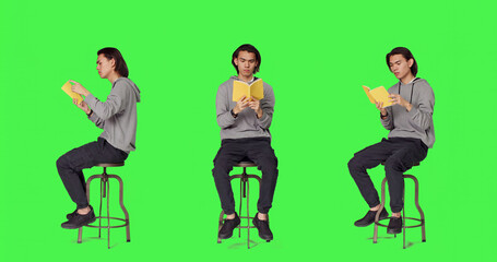 Obraz premium Trendy person reading novel book on chair against full body green screen template, enjoying old school lecture hobby, young asian adult holding literature or fiction book to read in studio