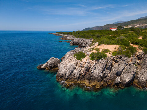 Aerial view of the rocky coastline of Aquarium Beach with turquoise waters and lush green vegetation under a clear blue sky in Jala, Vlore County, Albania.