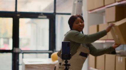 Fototapeta premium Warehouse advertising professional using smartphone to make branding video for social media, influencer filming herself in storage room showcasing company daily tasks to attract new employees.