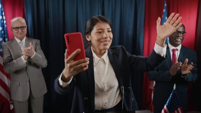 Young Hispanic woman politician live streaming on smartphone and waving at political rally while colleagues clap. Footage is suitable for election campaign promos