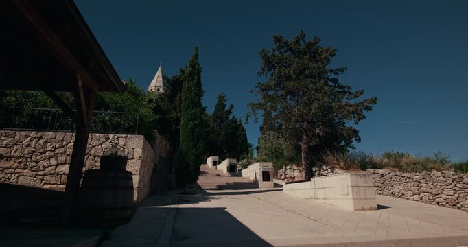 A paved path lined with trees and stone walls leads toward a church tower in the old town of Primosten, Croatia.