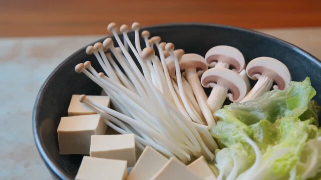 Fresh ingredients for hot pot including enoki mushrooms, button mushrooms, tofu cubes, and lettuce in a bowl.