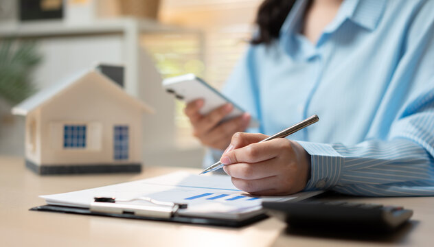 Businesswoman reviews financial documents with smartphone and pen. Her focused expression suggests careful planning. Miniature house model sits beside spreadsheets. Ideal for real estate, budgeting.