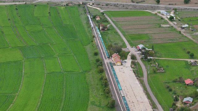 Top down aerial view of a passenger train passing by a small rural railway station next to vibrant green agricultural fields.