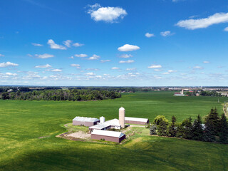 Low-level aerial drone flyover of a vibrant green corn field beside a rural road intersection, with farm buildings, silos, and open countryside stretching to the horizon. © Barry Lavallee