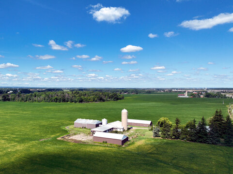 Low-level aerial drone flyover of a vibrant green corn field beside a rural road intersection, with farm buildings, silos, and open countryside stretching to the horizon.