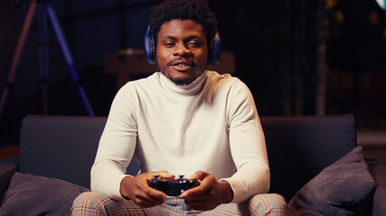 African American man having online gaming session with friends, talking with them through headphones microphone, gamer playing videogames with teammates on console using gamepad controller. © DC Studio