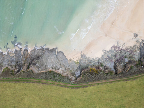 Aerial view of the rugged coastline featuring turquoise water, a sandy beach, rocky cliffs, and a coastal path bordering green fields in Padstow, Cornwall, United Kingdom.