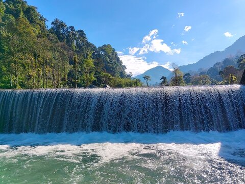 A horizontal landscape capturing water cascading over a river dam structure, creating a beautiful wide artificial waterfall surrounded by dense tropical forests and mountains on a bright sunny day.